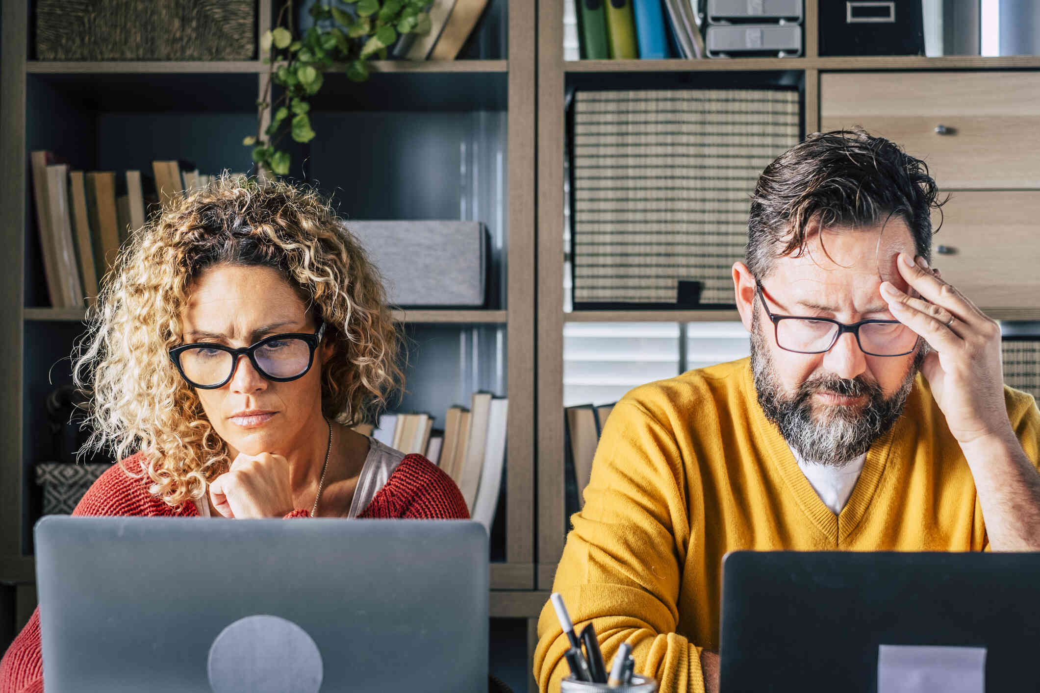 An older couple sits together, appearing stressed while both look at their own laptops.